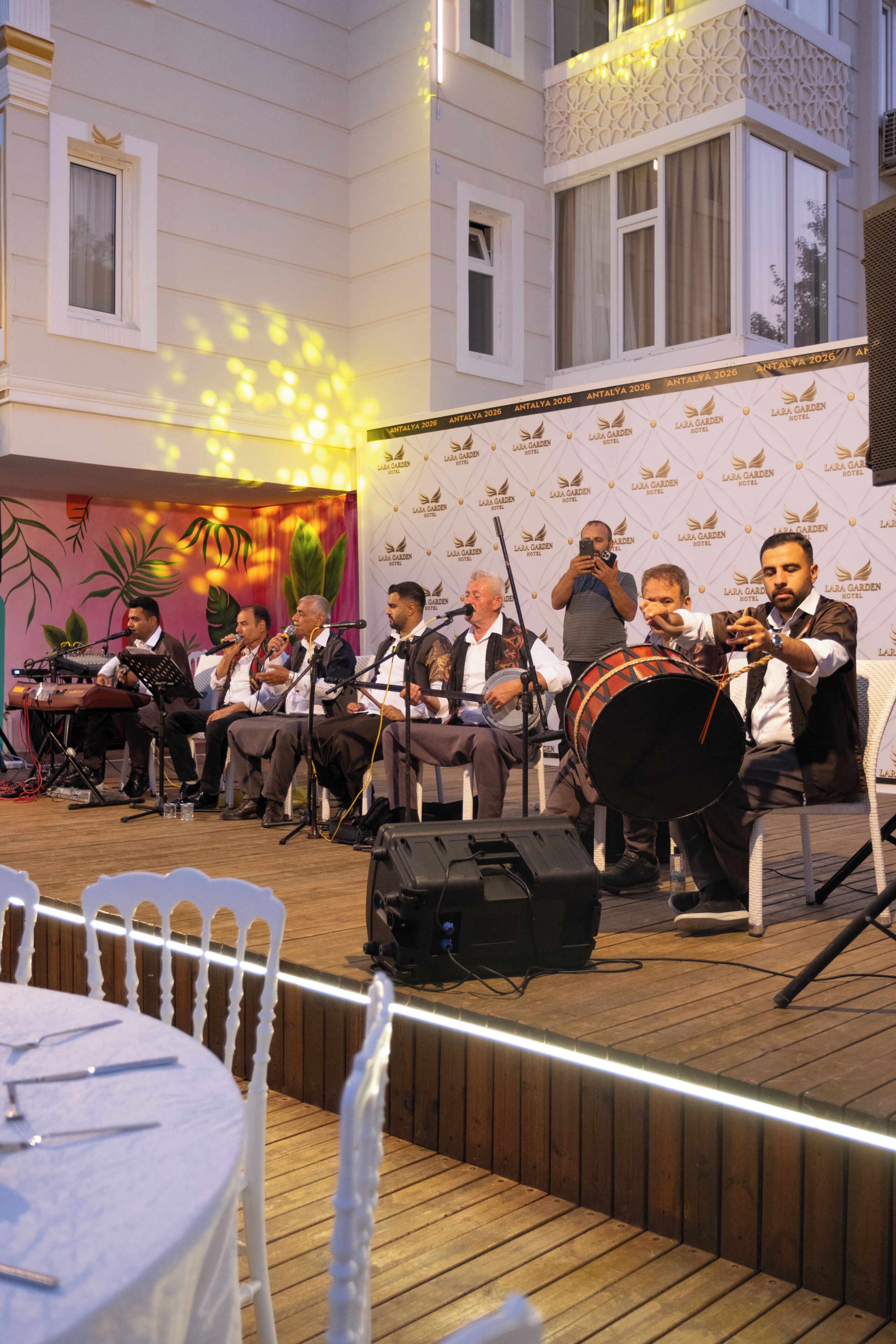 Una banda actuando en un escenario al aire libre con un fondo de diseño floral y el logo 'Lala Garden Hotel'.