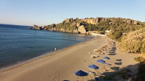 A beach with sun loungers and umbrellas, a clear sky, and rocky cliffs in the background. - Akrotiri Panorama (Agios Vasileios)