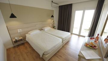 A hotel room with two beds, a bedside table with a phone and a lamp, a small table with a tray of fruit and drinks, and a window with curtains.