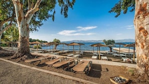 A beachside resort with sun loungers and umbrellas, a pool, and a view of the sea and mountains.