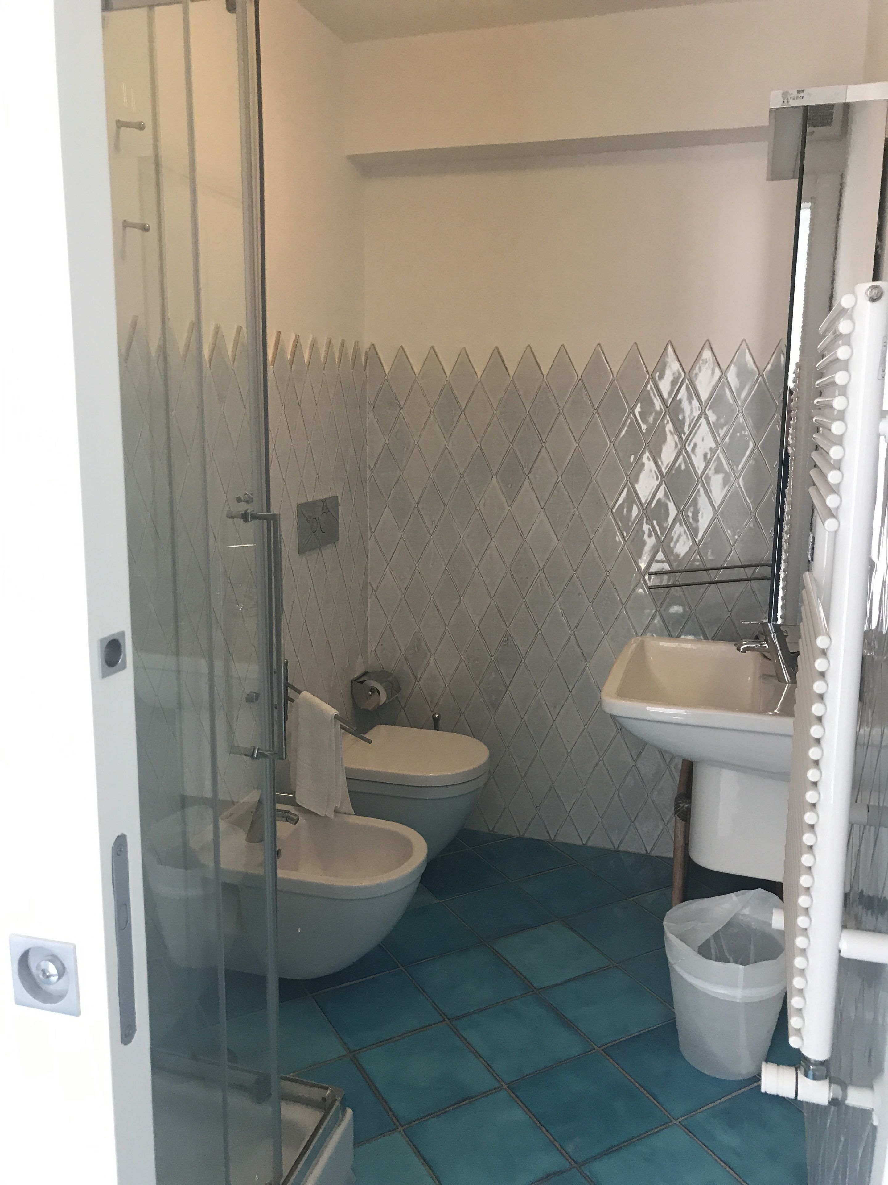 A bathroom with a glass shower enclosure, two toilets, a sink, and a trash bin.
