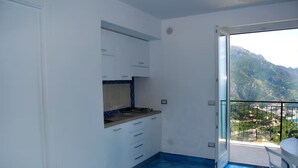 A kitchen with white cabinets and a blue-tiled floor, leading to a balcony with a view of mountains. - Residenza Sveva (Ravello)
