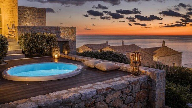 A rooftop pool area with stone walls, a hot tub, and a view of the sea at sunset. - Aria Estate (East Mani)