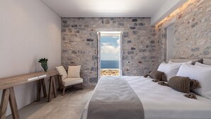 A bedroom with a stone wall, a bed with white linens, a wooden desk, a chair, and a view of the sea. - Aria Estate (East Mani)