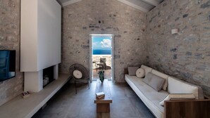 A stone-walled room with a high ceiling, a sofa, a chair, a small table, and a television. - Aria Estate (East Mani)