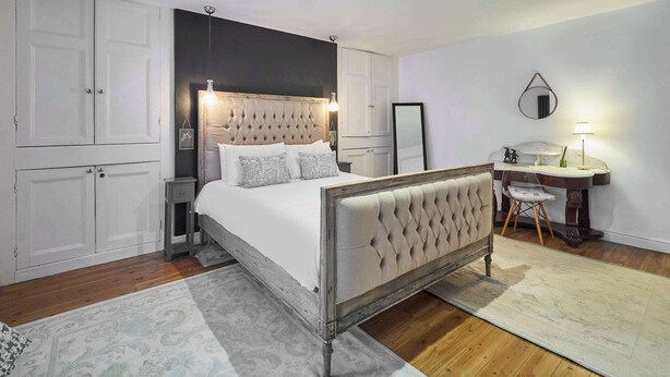 A bedroom with a large bed, a desk, and a chair. There is a wardrobe and a mirror on the wall.