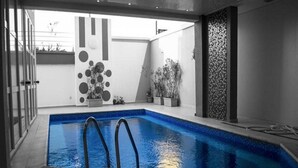 Piscina interior moderna com design geométrico na parede, vista clara do céu e plantas em vasos.