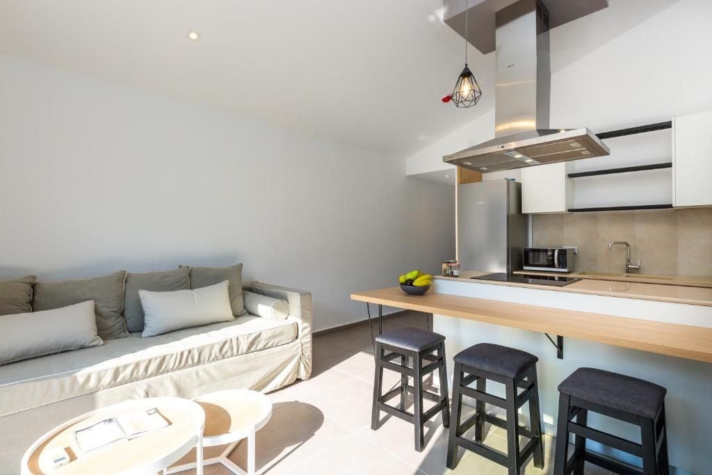 Un espacio habitable funcional con kitchenette, un sofá, una mesa de comedor y dos taburetes tipo bar.