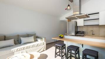 Un espacio habitable funcional con kitchenette, un sofá, una mesa de comedor y dos taburetes tipo bar.