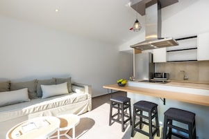Un espacio habitable funcional con kitchenette, un sofá, una mesa de comedor y dos taburetes tipo bar.