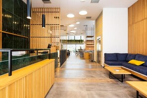 A modern reception area with a wooden reception desk, a blue sofa with a yellow pillow, and a kitchen in the background. - T Hotel Premium Suites (Mylopotamos)