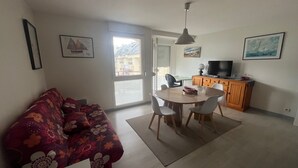 A living room with a sofa, dining table, chairs, a TV, and a kitchen area with cabinets and a window.