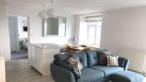 Uma sala de estar moderna com um sofá cinzento, uma área de jantar e uma cozinha ao fundo.