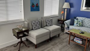 Una sala de estar con un sofá gris, un sillón a juego, una mesa de centro y una mesita de noche con una lámpara. Hay dos ventanas con persianas, un reloj en la pared y un cuadro sobre el sofá.