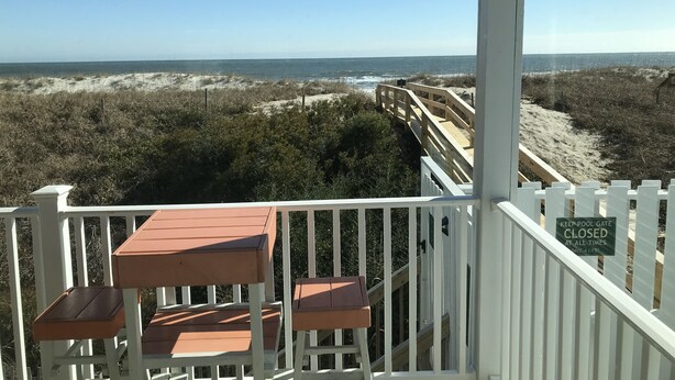 Vista a la playa desde un porche cubierto con mesas de madera y una cerca blanca de piquete.