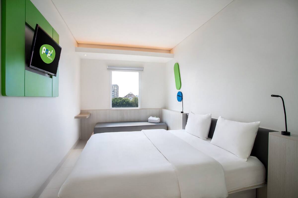 A modern hotel room with a large bed, a flat-screen TV mounted on a green panel, a window with a view of buildings, and a small seating area.