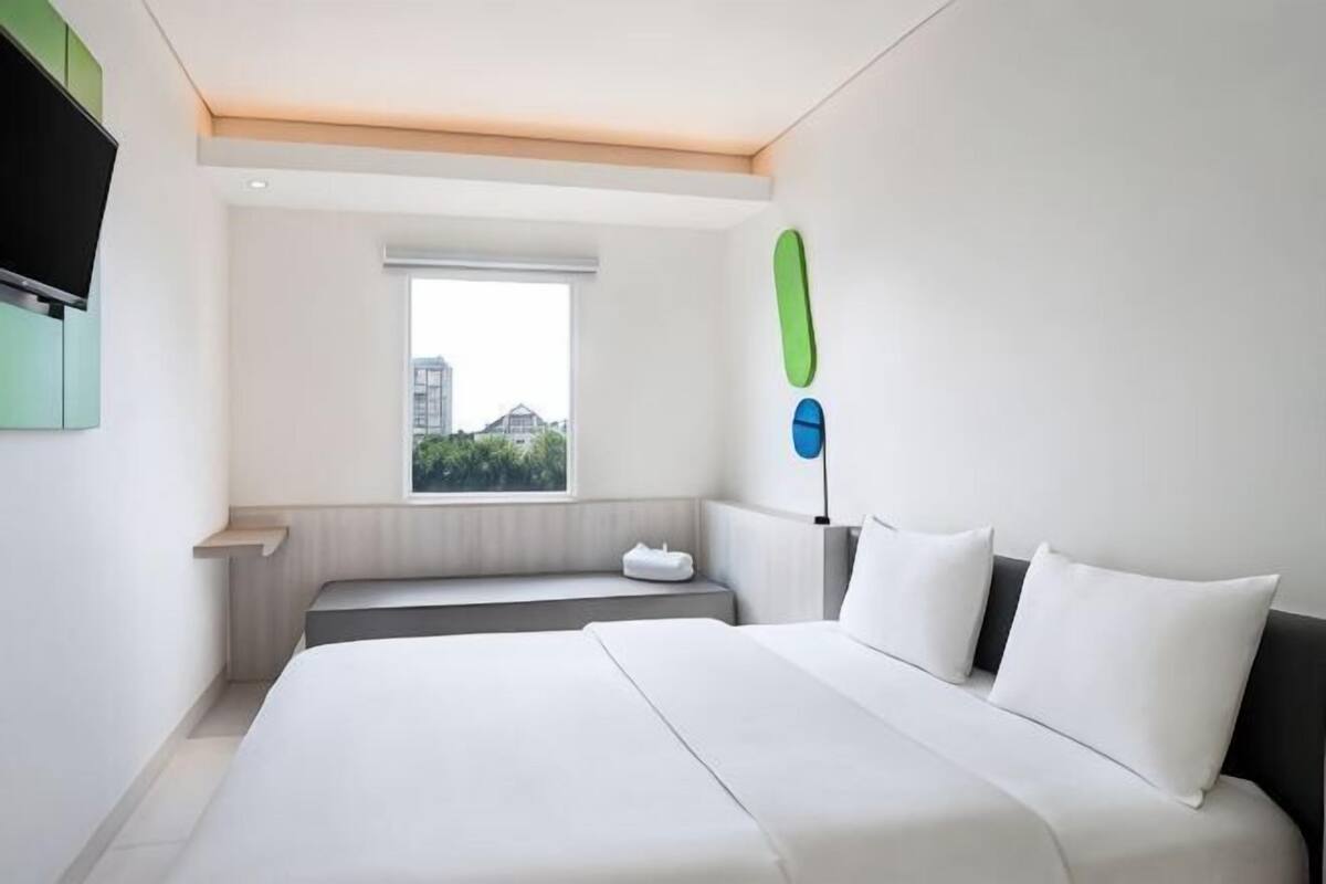 A modern hotel room with a large bed, a window with a city view, a flat-screen TV, and a small seating area.