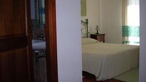 A bedroom with a bed, a nightstand, a lamp, a window with curtains, and a bathroom with a sink and mirror. - Bed and Breakfast Dessole (Olbia)