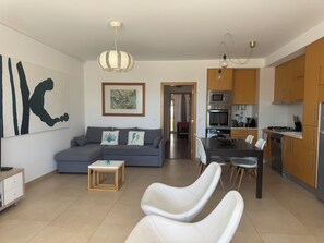 A modern living room with a grey sofa, a small wooden table, and a dining area with white chairs. A kitchen with wooden cabinets and stainless steel appliances is visible in the background.
