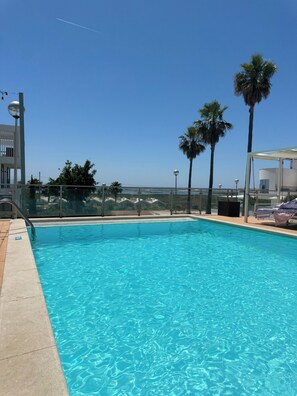 A clear blue pool with a view of the ocean, palm trees, and a modern building structure.