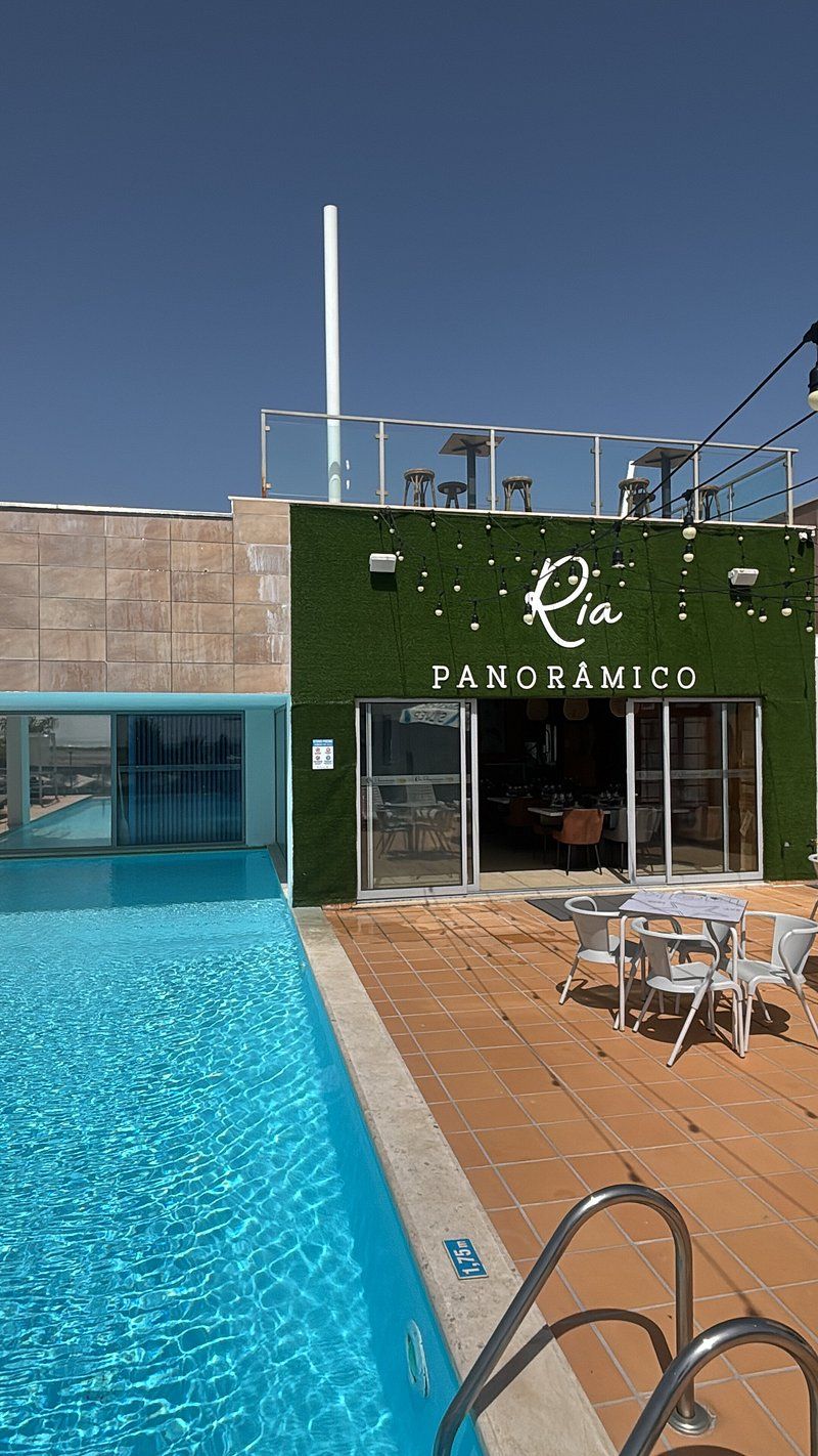 A swimming pool with a clear blue water, a restaurant named 'Ria Panoramico' with outdoor seating, and a tiled deck area.