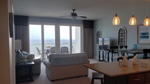 A living room with a sofa, a dining table, and a TV. There are curtains, a ceiling fan, and a view of the ocean. - Lake Front-Private Beach Access- Golf - all within walking distance (Panama City)