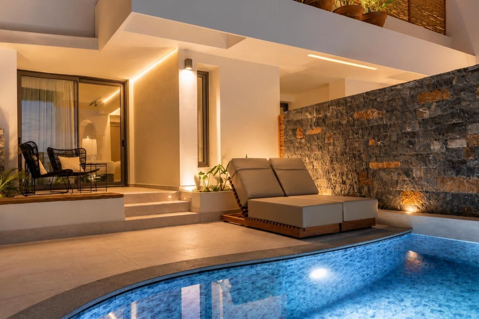 Ein moderner, überdachter Poolbereich mit Steinwand, einer Sitzecke mit Kissen und einer Glastür zu einem weiteren Raum.