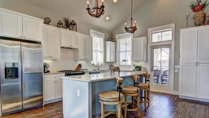 Une cuisine moderne avec des meubles blancs, un réfrigérateur en acier inoxydable, un îlot central doté de tabourets de bar et un éclairage décoratif.