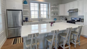 A modern kitchen with white cabinetry, a stainless steel refrigerator, a large island with bar stools, and a window providing natural light. - Cinnamon Shore- Sea Change 115 FI (Port Aransas)