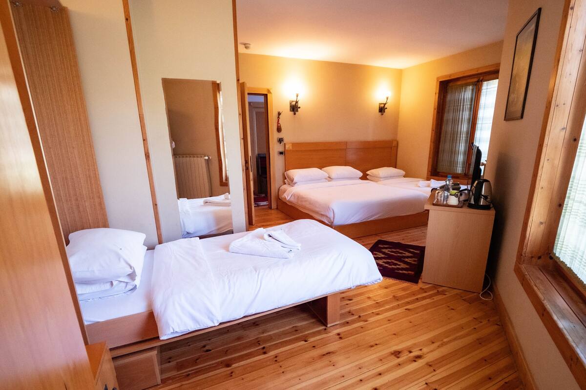 A hotel room with two beds, wooden flooring, a desk with a kettle, and a window with curtains.