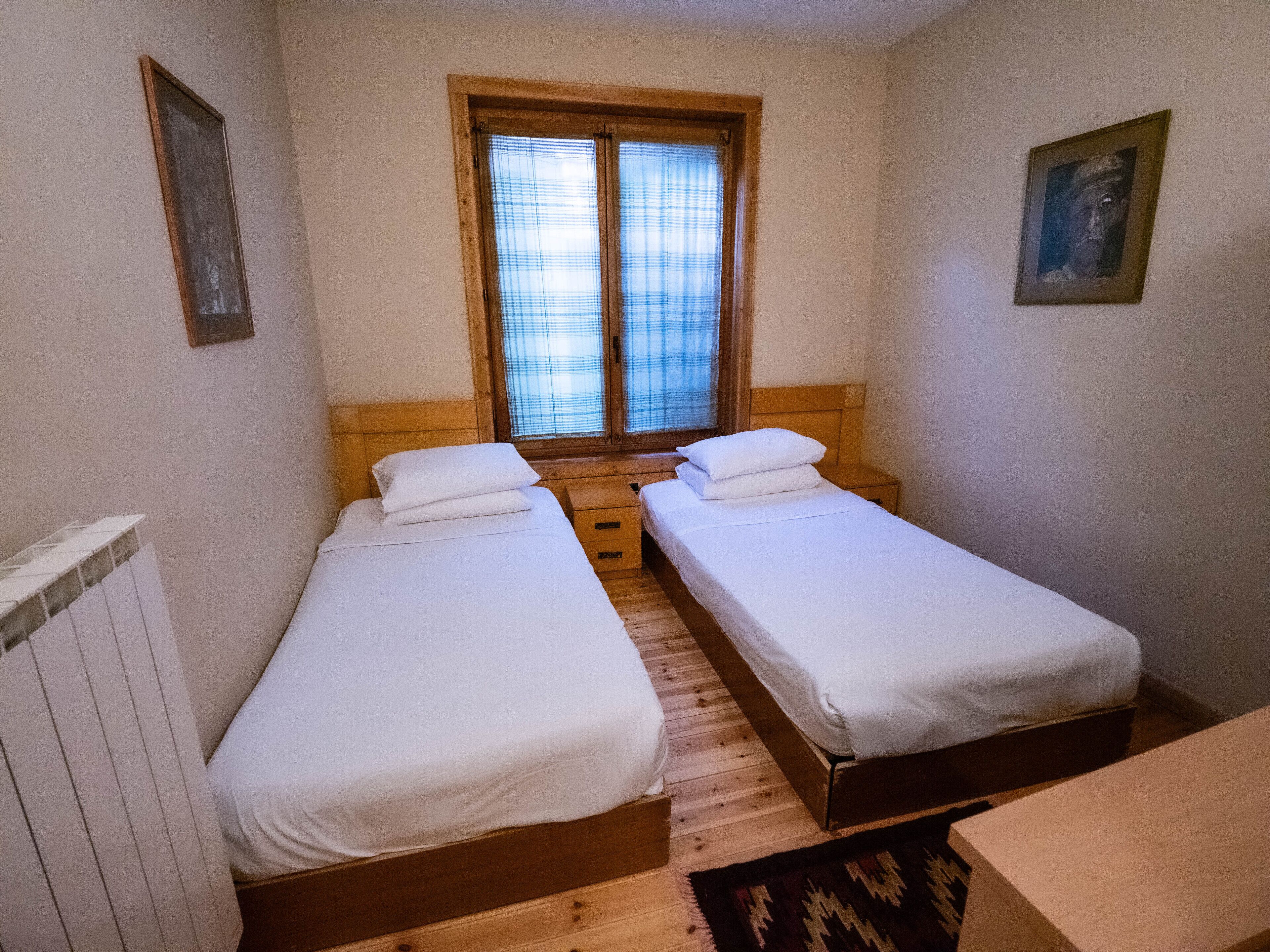 Ein kleines, sauberes Zimmer mit zwei Einzelbetten, Holzboden, einem Fenster mit Jalousien und einer Heizung.