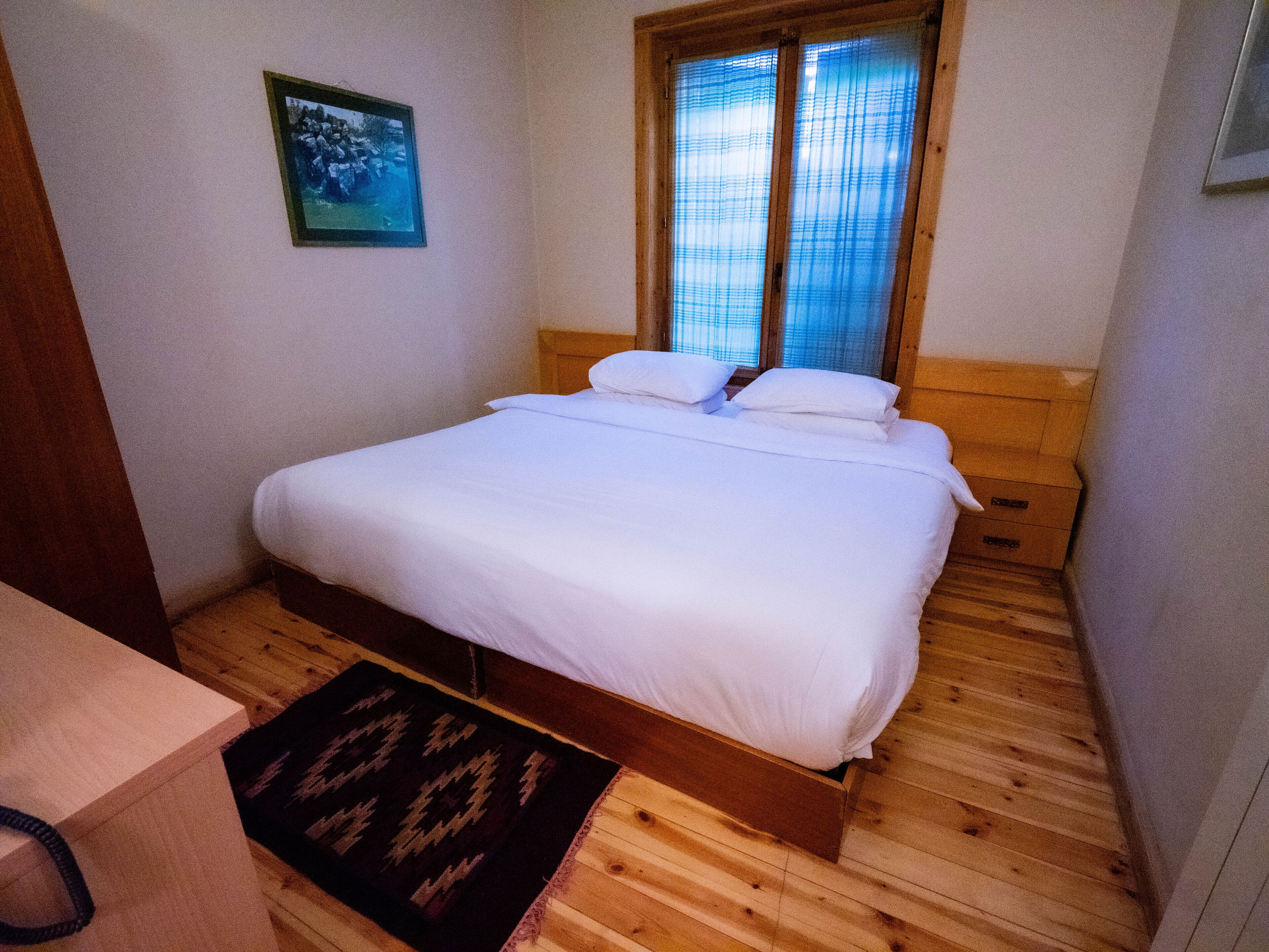 Un dormitorio con cama de madera, una ventana con persianas, un cuadro enmarcado en la pared y una pequeña alfombra en el suelo.