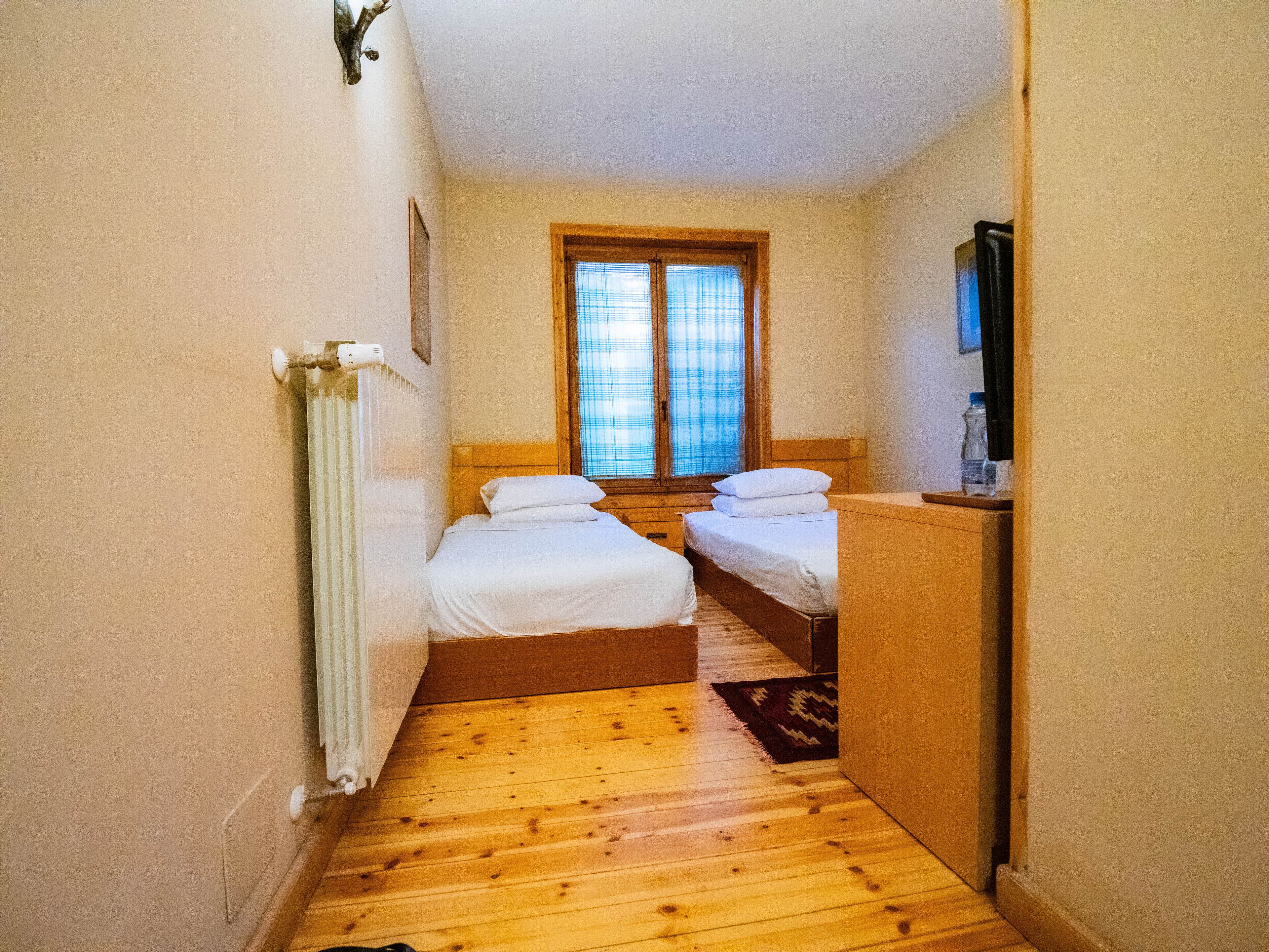 Una habitación pequeña con dos camas individuales, un radiador, una ventana con persianas y un televisor sobre un mueble de madera.