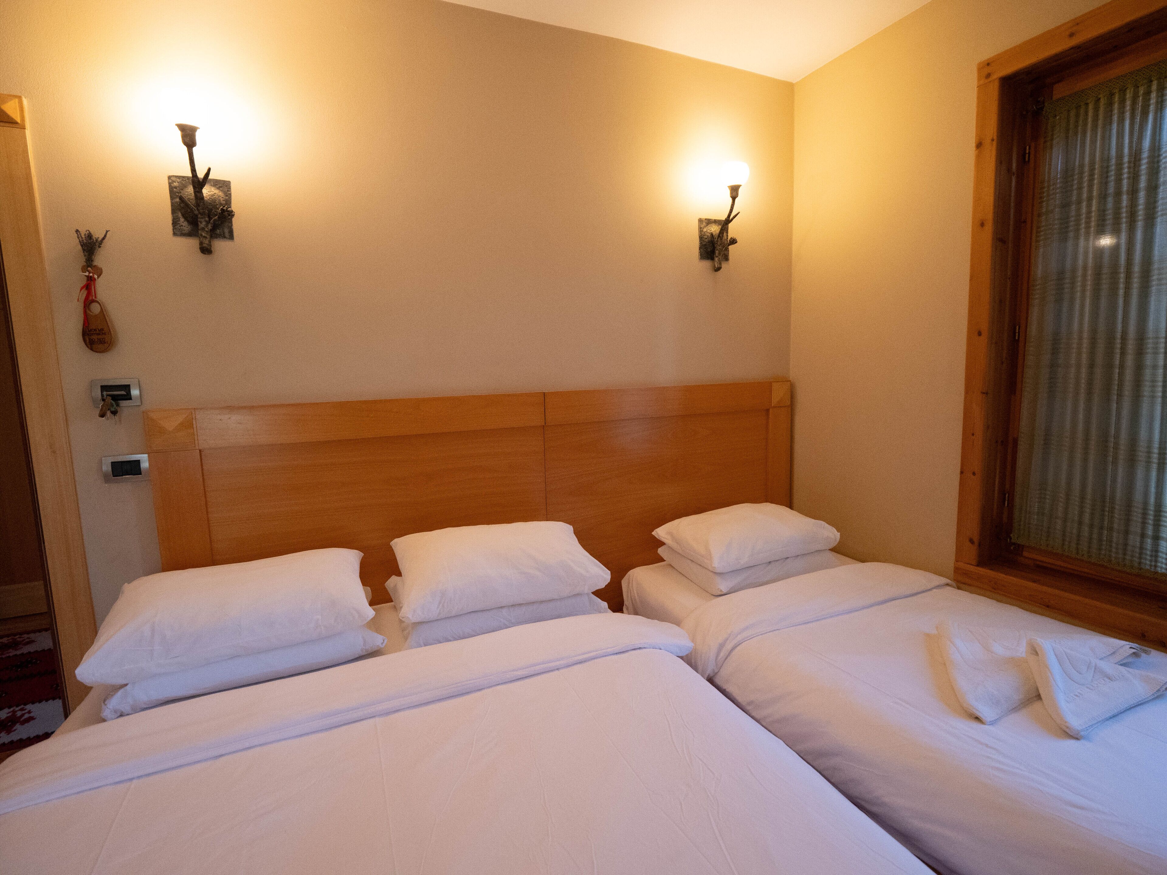 Hotellihuone, jossa on kolme sänkyä, puiset päätyosat, seinään kiinnitetyt valaisimet ja ikkuna, jossa on verhot.