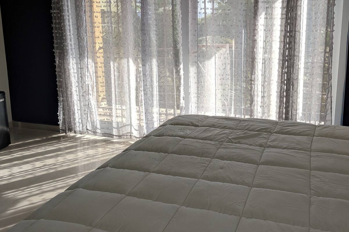 A bed with a quilted cover in a room with sheer curtains and a view of the outside.