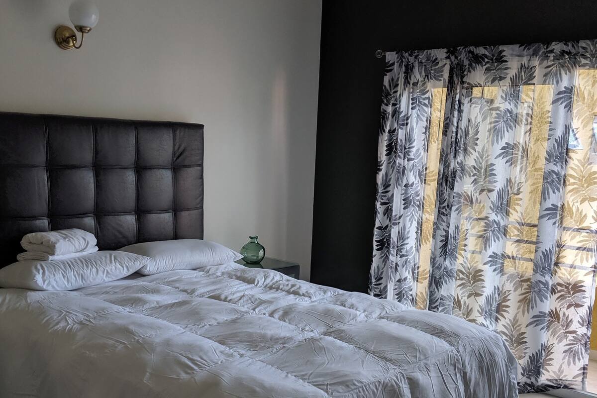 A neatly made bed with a dark headboard, a bedside table with a green vase, and a window with floral curtains.