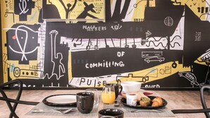 A breakfast table set with plates, mugs, and a glass of orange juice, with a mural in the background. - Arena Hotel La Defense (Nanterre)