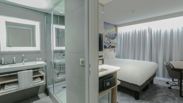 A modern hotel room with a glass-enclosed shower, a large bed, a desk, and a television. - Arena Hotel La Defense (Nanterre)