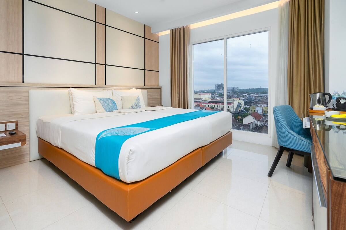 A hotel room with a large bed, a desk with a chair, and a view of the city through large windows.