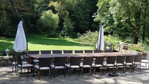 Une table de jardin rectangulaire, conçue pour accueillir huit personnes, entourée de gravier, et située dans un jardin agrémenté d’arbres et d’un pont en bois en arrière-plan.