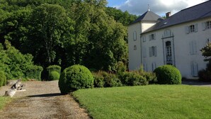 Un grand bâtiment blanc, surmonté d’un toit gris, entouré d’une végétation luxuriante et accessible par un chemin de gravier menant à l’entrée.