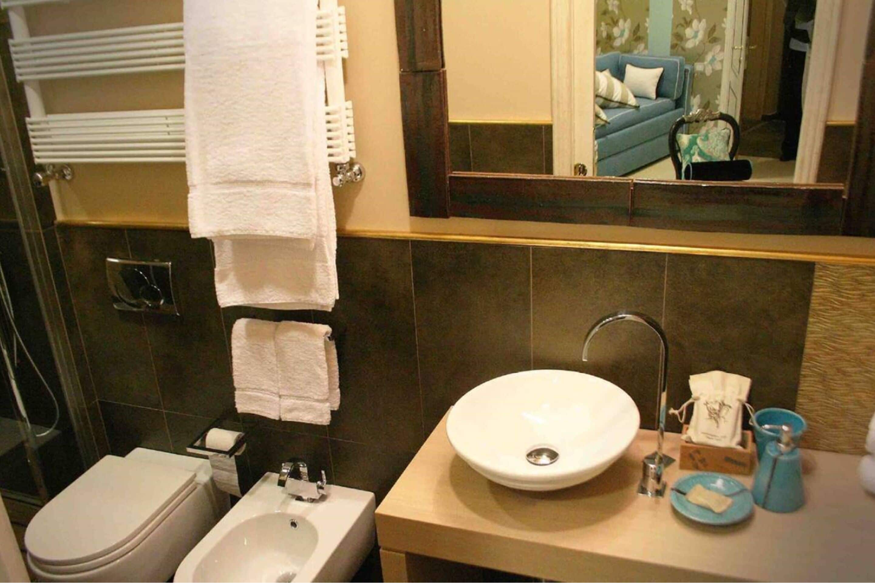 A bathroom with a toilet, bidet, sink, and a mirror reflecting a living room with a sofa and a coffee table.