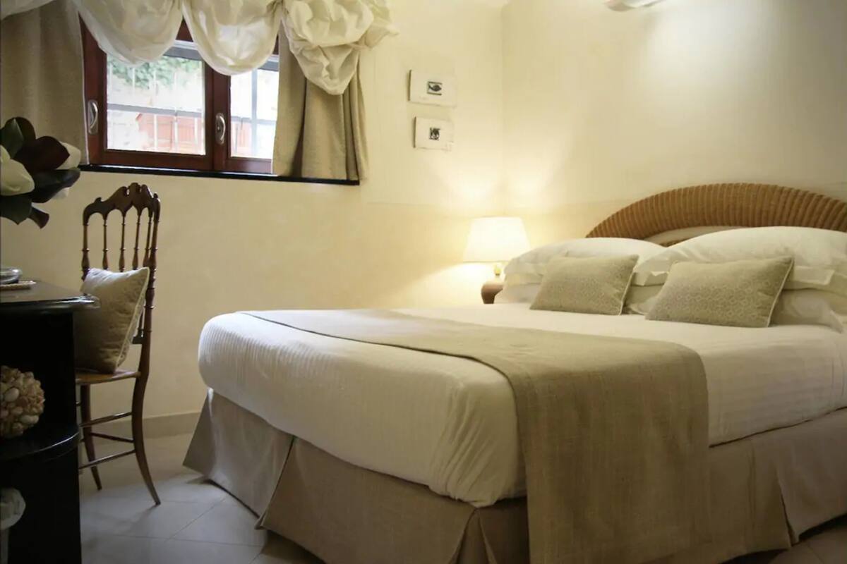 A bedroom with a large bed, a wooden chair, a desk, and a window with curtains.