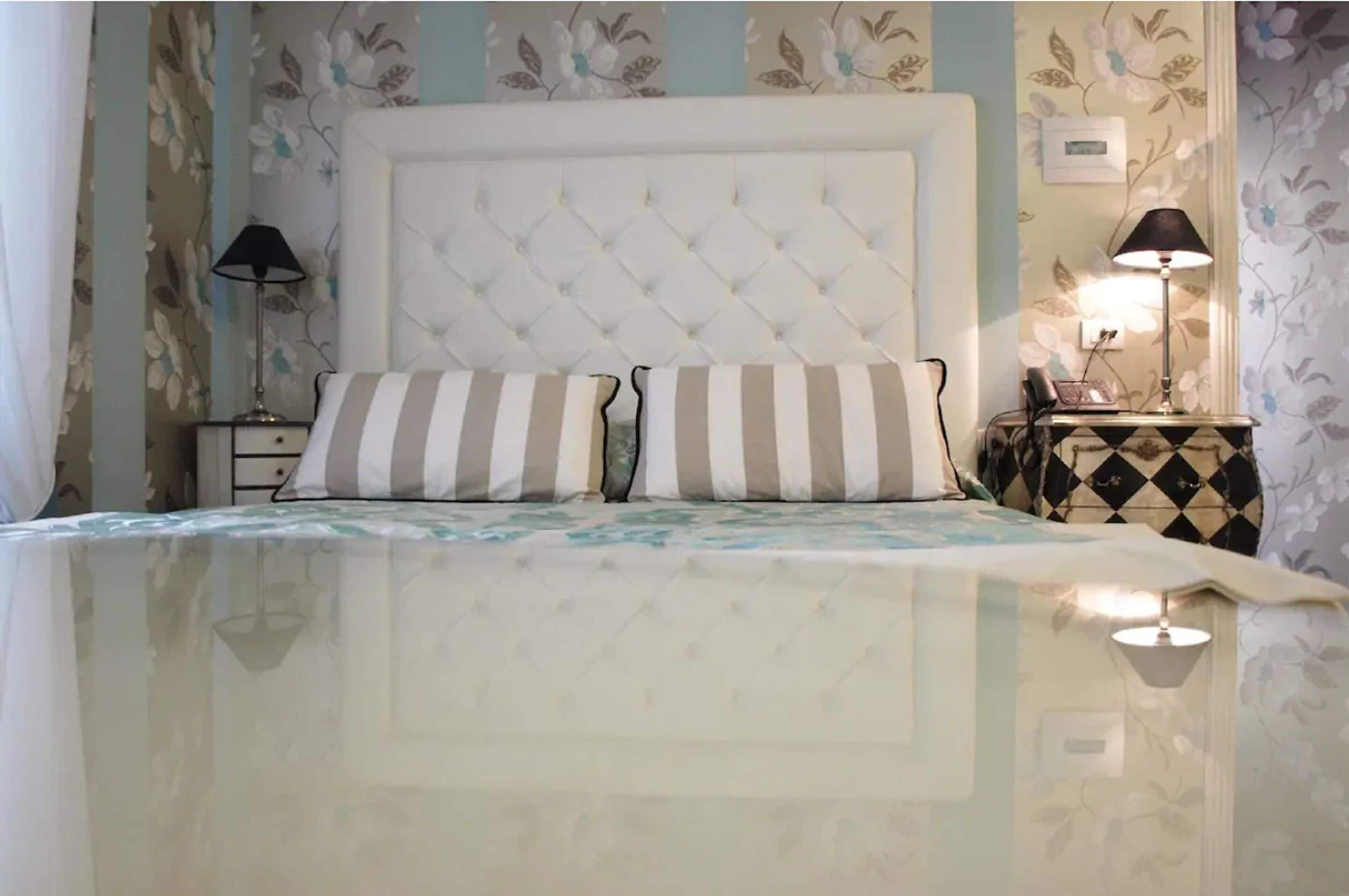 Un dormitorio con cabecera capitonada, almohadas a rayas, una mesita de noche con lámpara y papel tapiz floral.