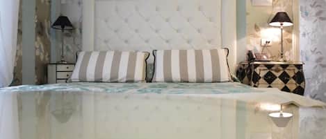 A bedroom with a tufted headboard, striped pillows, a bedside table with a lamp, and floral wallpaper.