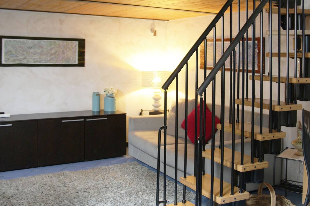 A staircase with a wooden finish and black metal railings, leading to an upper level. A white sofa with a red pillow is positioned below the stairs.