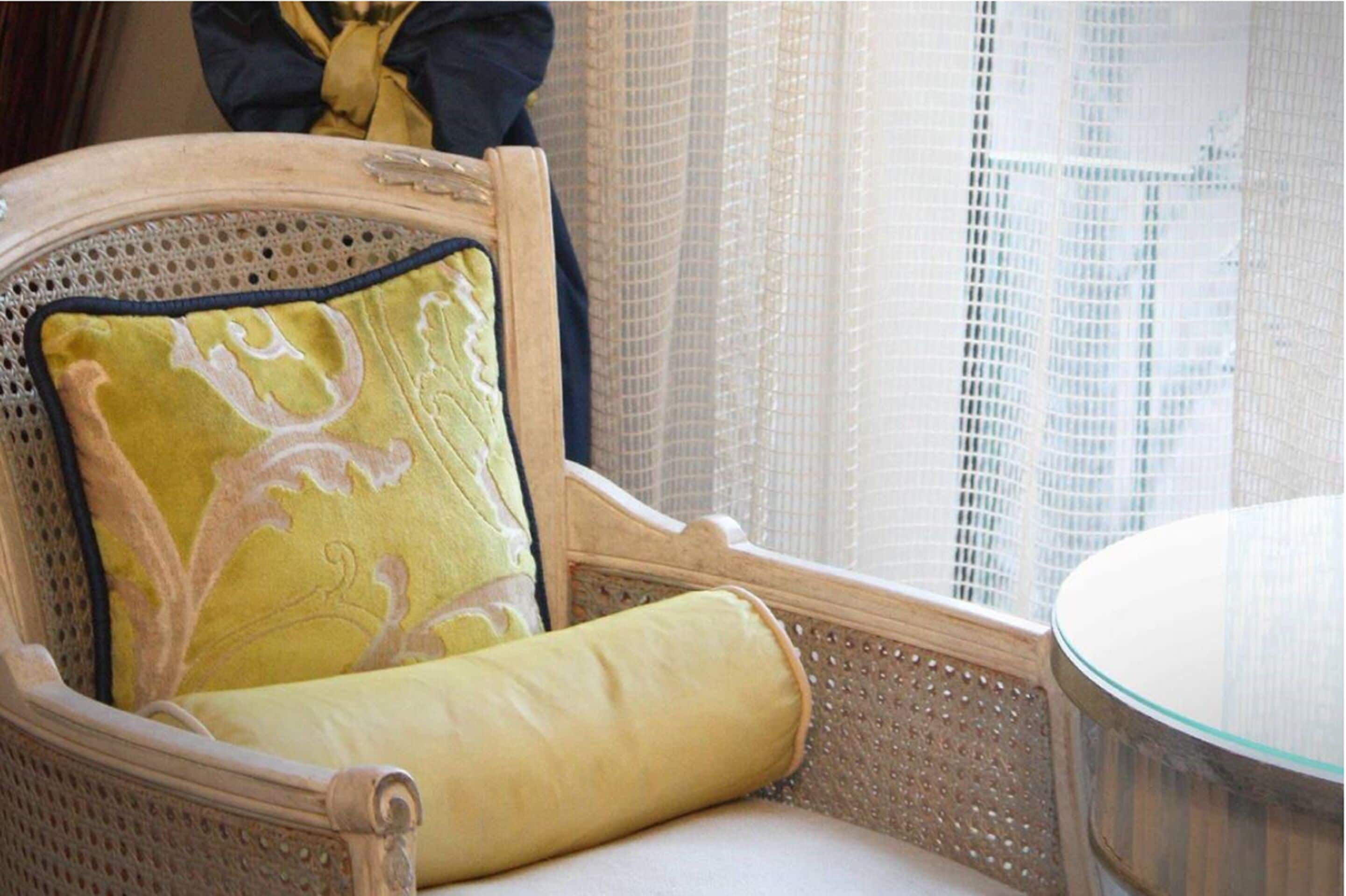 A chair with a decorative pillow and a cushion, a glass table, and a window with sheer curtains.