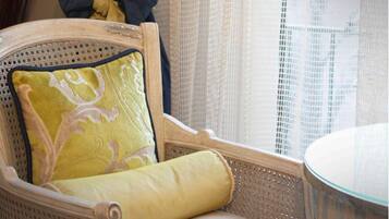 A chair with a decorative pillow and a cushion, a glass table, and a window with sheer curtains.