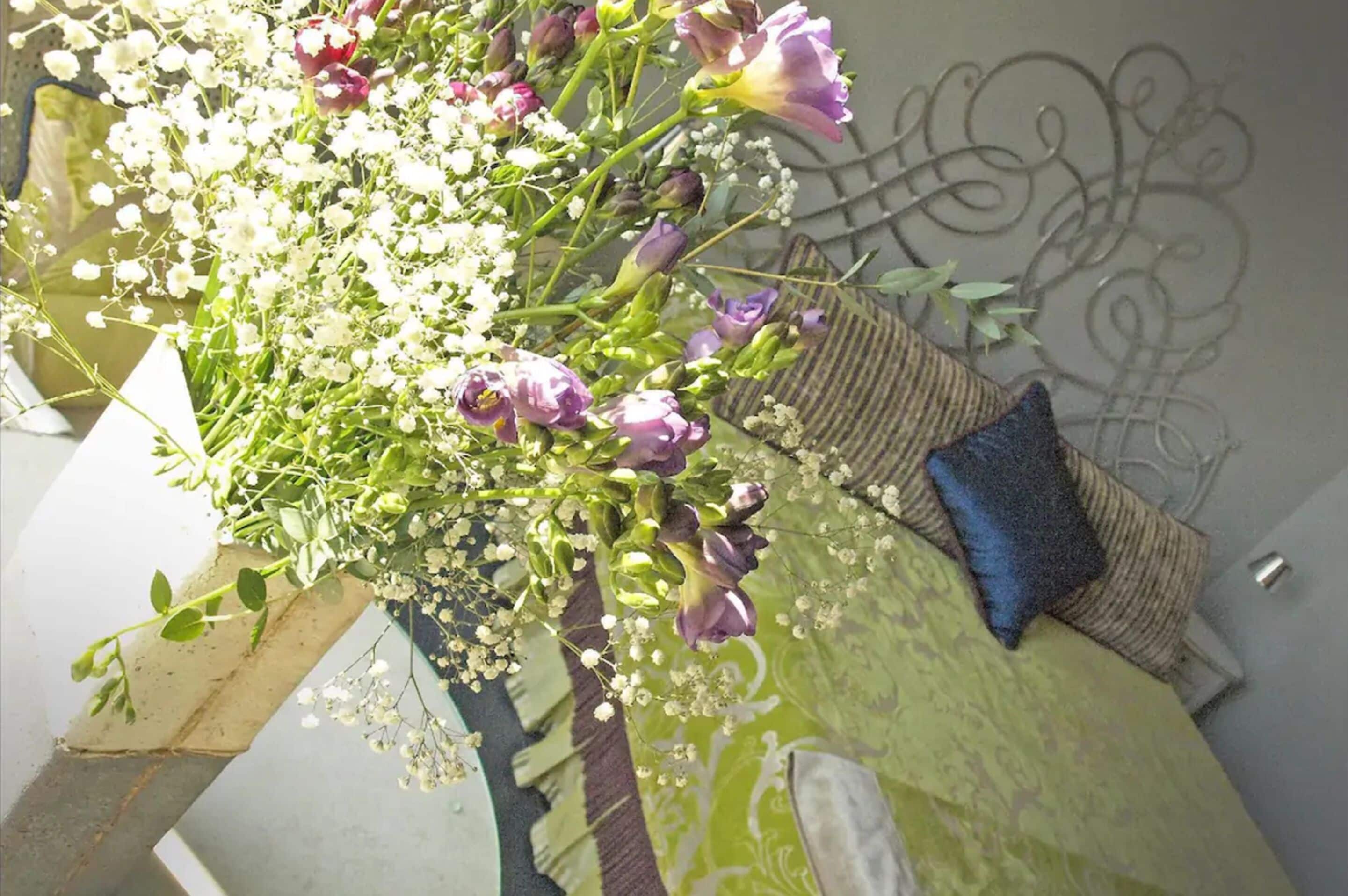 Um arranjo floral num canto do quarto, com uma almofada estampada e um ornamento metálico na parede.