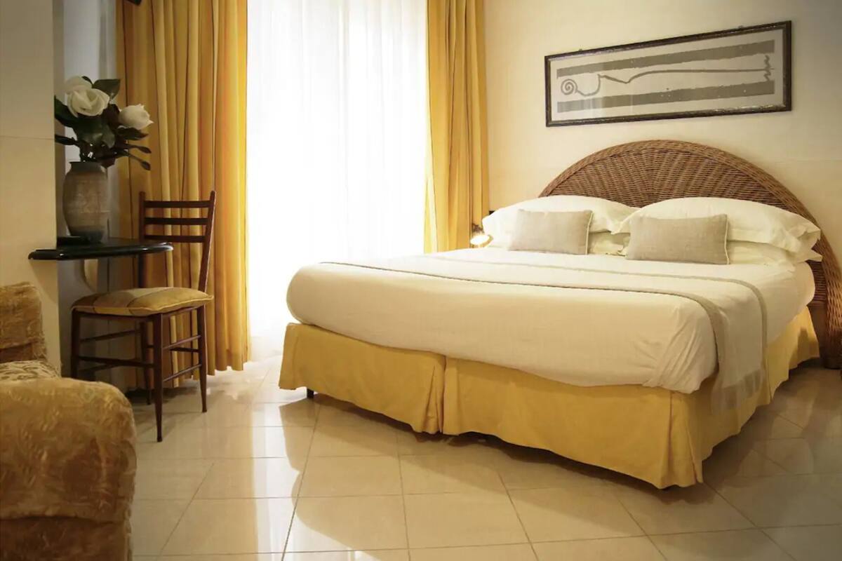 A neatly made bed with a yellow bed skirt, a chair, and a vase with flowers on a small table.